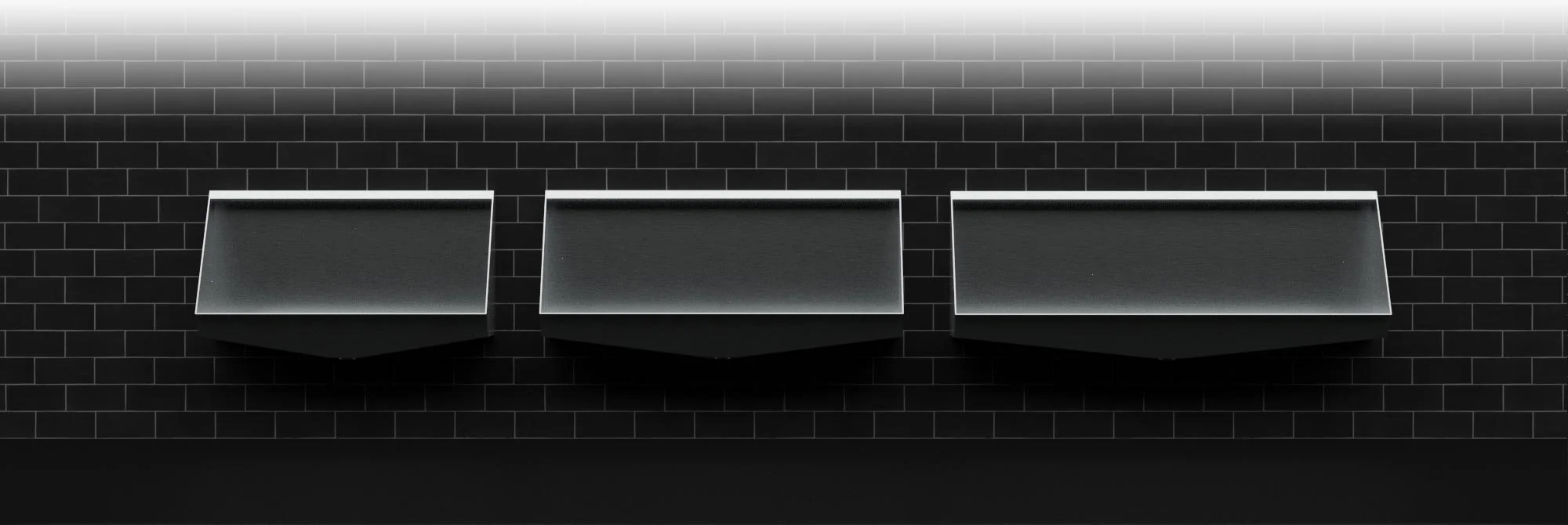 A selection of stainless steel urinal troughs in varying sizes fixed to dark coloured tiled wall in a washroom